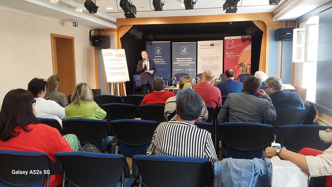 Sala konferencyjna z widownią skierowaną w stronę sceny. Na pierwszym planie znajdują się rzędy niebieskich krzeseł, na których siedzą kobiety i mężczyźni w różnym wieku, ubrani w kolorowe ubrania: czerwień, zieleń, biel, szarość, granat. Na scenie pośrodku stoi mężczyzna w ciemnym garniturze z mikrofonem w dłoni i kartkami, wygląda, jakby prowadził wystąpienie. Za nim ustawione są trzy ciemnoniebieskie roll‑upy z białymi napisami, między innymi „INSTYTUT HISTORYCZNY” oraz „MIĘDZYNARODOWY OŚRODEK BADAŃ INTERDYSCYPLINARNYCH W SŁUŻBIE EDUKACJI”. Obok nich widoczny jest biały roll‑up z logotypami i tekstem oraz czerwono‑pomarańczowy roll‑up z napisem „Trzymaj się Granicy! Szlakiem architektury obronnej”. Z lewej strony sceny stoi tablica na sztaludze z wydrukowanym tekstem, po prawej stronie sceny stoi kobieta w zielonej sukience przy stoliku. Ściany są jasne, a nad sceną i nad widownią zawieszony jest sprzęt oświetleniowy. Po prawej stronie kadru znajduje się okno z otwartymi skrzydłami, wpuszczające światło dzienne.