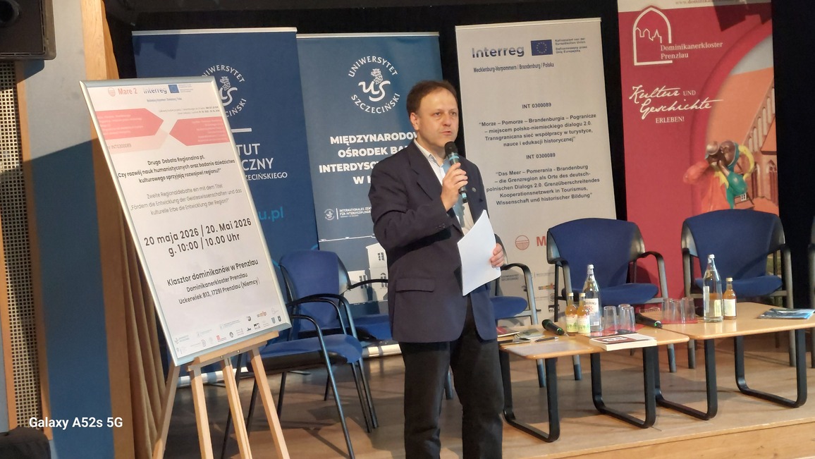 Mężczyzna w garniturze stojący na scenie z mikrofonem. Mężczyzna o jasnej skórze, w ciemnej marynarce, jasnej koszuli i ciemnych spodniach stoi na drewnianej podłodze, trzyma mikrofon w prawej ręce i kilka kartek w lewej, wygląda jak prowadzący lub prelegent podczas konferencji. Za nim ustawione są trzy duże pionowe banery z logotypami: po lewej granatowy z napisem „Uniwersytet Szczeciński” i „Międzynarodowy Ośrodek Interdyscyplinarny”, pośrodku jasny z logotypami Unii Europejskiej i programu Interreg, po prawej czerwony z napisem „Dominikanerkloster Prenzlau” oraz grafiką przedstawiającą postać w kolorowych ubraniach. Po lewej stronie zdjęcia stoi tablica na sztaludze z dużym afiszem informacyjnym; na plakacie widoczna jest m.in. data „20 maja 2026 / 20. Mai 2026, g. 10:00 / 10.00 Uhr” oraz miejsce „Klasztor dominikanów w Prenzlau, Uckerwiek 813, 17291 Prenzlau (Niemcy)”. Na pierwszym planie po prawej stronie znajduje się niski prostokątny stolik, na którym stoją butelki z napojami, plastikowe kubki oraz kilka broszur lub czasopism; za stolikiem ustawione są niebieskie krzesła konferencyjne.