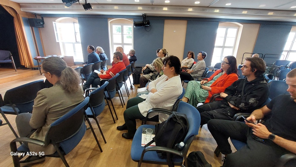 Sala konferencyjna z kilkunastoma osobami siedzącymi w rzędach krzeseł, skierowanymi w stronę sceny po lewej stronie. Widziana jest część małej sceny z drewnianą podłogą i zasłonami w kolorze złoto‑żółtym po lewej. Krzesła są głównie niebieskie z czarnymi podłokietnikami, ustawione w kilku rzędach. Osoby siedzą w większości bokiem, zwrócone w stronę sceny, wyglądają jak uczestnicy wykładu lub prezentacji, część z nich ma założone ręce, część coś notuje lub trzyma telefony. Uczestnicy są w różnym wieku, od młodych dorosłych do osób starszych, w strojach od bardzo casualowych po bardziej eleganckie, z kilkoma wyraźnymi kolorami, jak czerwone marynarki i zielona sukienka w jasny wzór. W tle po prawej stronie ściana ma kilka dużych okien o białych ramach, przez które wpada jasne światło dzienne. Na suficie znajdują się oprawy oświetleniowe i głośniki, a pośrodku ściany z oknami zamontowany jest czarny głośnik lub projektor na wysięgniku. Na pierwszym planie przy jednym z krzeseł stoi mały stolik lub podłokietnik z plastikowym kubkiem i notatkami.