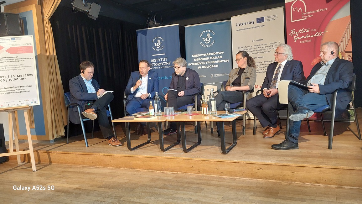 Panel dyskusyjny na scenie z sześcioma osobami siedzącymi na krzesłach w półokręgu. Na środku stoi prostokątny stolik z jasnym blatem i ciemnymi metalowymi nogami; na blacie leżą dokumenty, książki, kilka plastikowych kubków i dwie szklane butelki z napojami. Za panelistami ustawiono pięć dużych stojących roll‑upów i plansz z logotypami, tekstami po polsku i niemiecku oraz informacjami o instytucjach: po lewej jasna tablica informacyjna na sztaludze, obok niej dwa granatowe banery Uniwersytetu Szczecińskiego i Instytutu Historycznego, dalej biały baner projektu Interreg oraz po prawej czerwony baner Dominikanerkloster Prenzlau. Od lewej siedzi mężczyzna o jasnej karnacji w ciemnej marynarce, jasnej koszuli i ciemnych spodniach, trzyma notatnik na kolanach; obok mężczyzna w granatowym garniturze z mikrofonem w dłoni, pochylony lekko do przodu; następnie mężczyzna z siwymi włosami i brodą, w ciemnym garniturze bez krawata, patrzący w dół na dokument; obok kobieta o jasnej karnacji, z długimi włosami związanymi z tyłu, w ciemnych dżinsach i koszulce oraz krótkiej kurtce, siedząca z nogą założoną na nogę; dalej mężczyzna z siwymi włosami i wąsami, w granatowym garniturze, białej koszuli i wzorzystym krawacie, trzymający dłonie splecione na kolanie; po prawej skrajnej stronie mężczyzna o jasnej karnacji w ciemnej marynarce i niebieskiej koszuli, z założoną nogą i słuchawką w uchu, trzymający kartki. Scena ma drewnianą podłogę, czarne tło i z lewej strony beżową kurtynę; nad sceną widoczne są reflektory oświetlające uczestników.