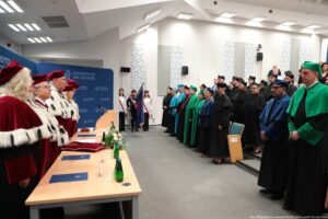 Uroczysta akademicka ceremonia w dużej auli. Po lewej stronie przy długim stole stoją osoby w bordowych togach z białym, futrzanym obszyciem i bordowych biretach, wyglądające na rektorów lub władze uczelni. Na stole leżą granatowe teczki, ciemnoczerwone poduszki, są butelki z wodą i bukiet kwiatów. Za stołem widoczna jest niebieska ścianka z białym logo i napisem „UNIWERSYTET SZCZECIŃSKI”. Na wprost, po prawej stronie zdjęcia, stoi rząd osób w togach w różnych kolorach: zielonych, niebieskich, turkusowych oraz czarnych, z dopasowanymi biretami. W tle, pośrodku, stoją trzy osoby z pocztem sztandarowym: trzymają flagę, mają biało‑czerwone szarfy oraz bordowe nakrycia głowy. W dalszych rzędach siedzą lub stoją inni uczestnicy w czarnych togach. Pomieszczenie jest jasne, z białymi ścianami, kilkoma panelami akustycznymi i prostokątnymi sufitowymi lampami. Cała scena sprawia wrażenie formalnej inauguracji lub innej ważnej uroczystości akademickiej.