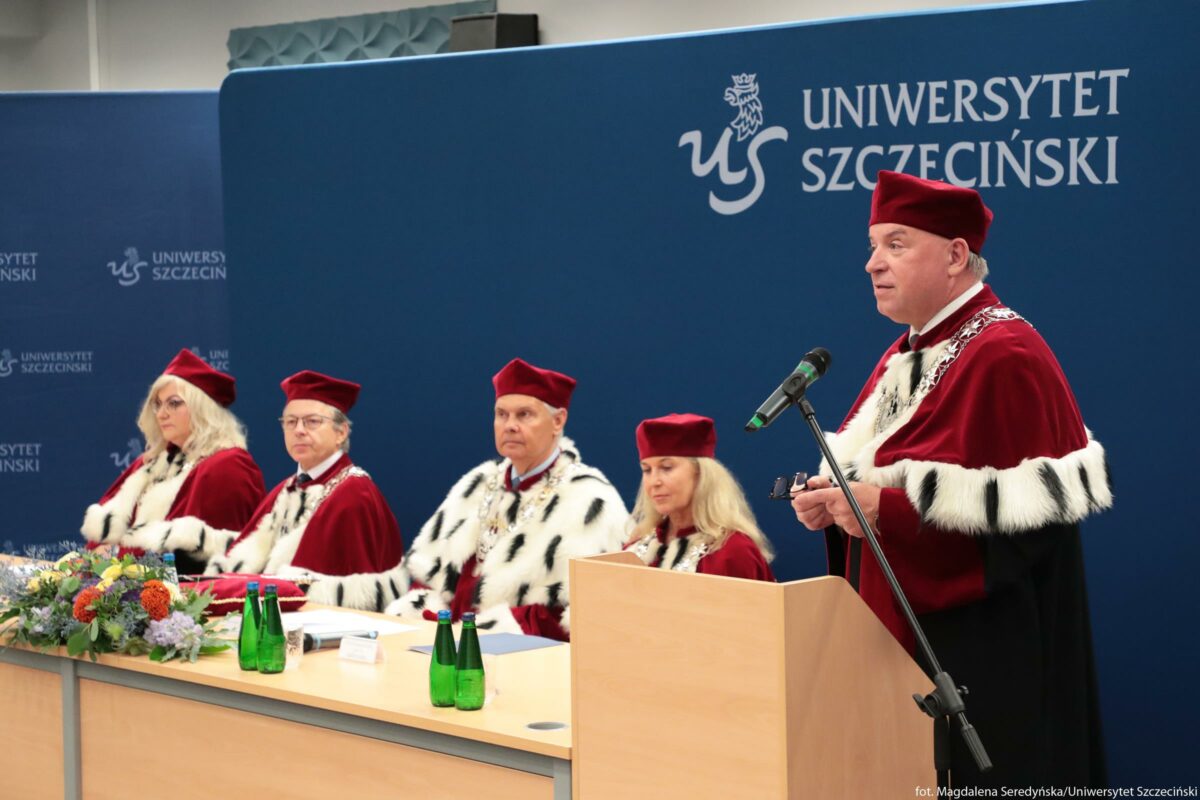 Grupa pięciu osób w uroczystych togach siedząca i stojąca przy stole na tle niebieskiej ściany. Wszyscy mają bordowe aksamitne birety i czerwone togi obszyte białym futrem w czarne cętki, przypominające tradycyjne stroje akademickie. Po prawej stronie mężczyzna o jasnej skórze stoi za drewnianą mównicą, trzyma w dłoni mały przedmiot i mówi do mikrofonu na statywie. Po lewej stronie cztery osoby siedzą za długim stołem: kobieta o jasnych, długich włosach, dwóch mężczyzn oraz kolejna kobieta o jasnych włosach; wszyscy mają na szyi łańcuchy rektorskie lub dziekańskie. Na stole stoją zielone szklane butelki z wodą, leżą dokumenty oraz bukiet kolorowych kwiatów. W tle na niebieskim panelu widoczny jest biały napis „UNIWERSYTET SZCZECIŃSKI” oraz biały znak graficzny z literami „US”.