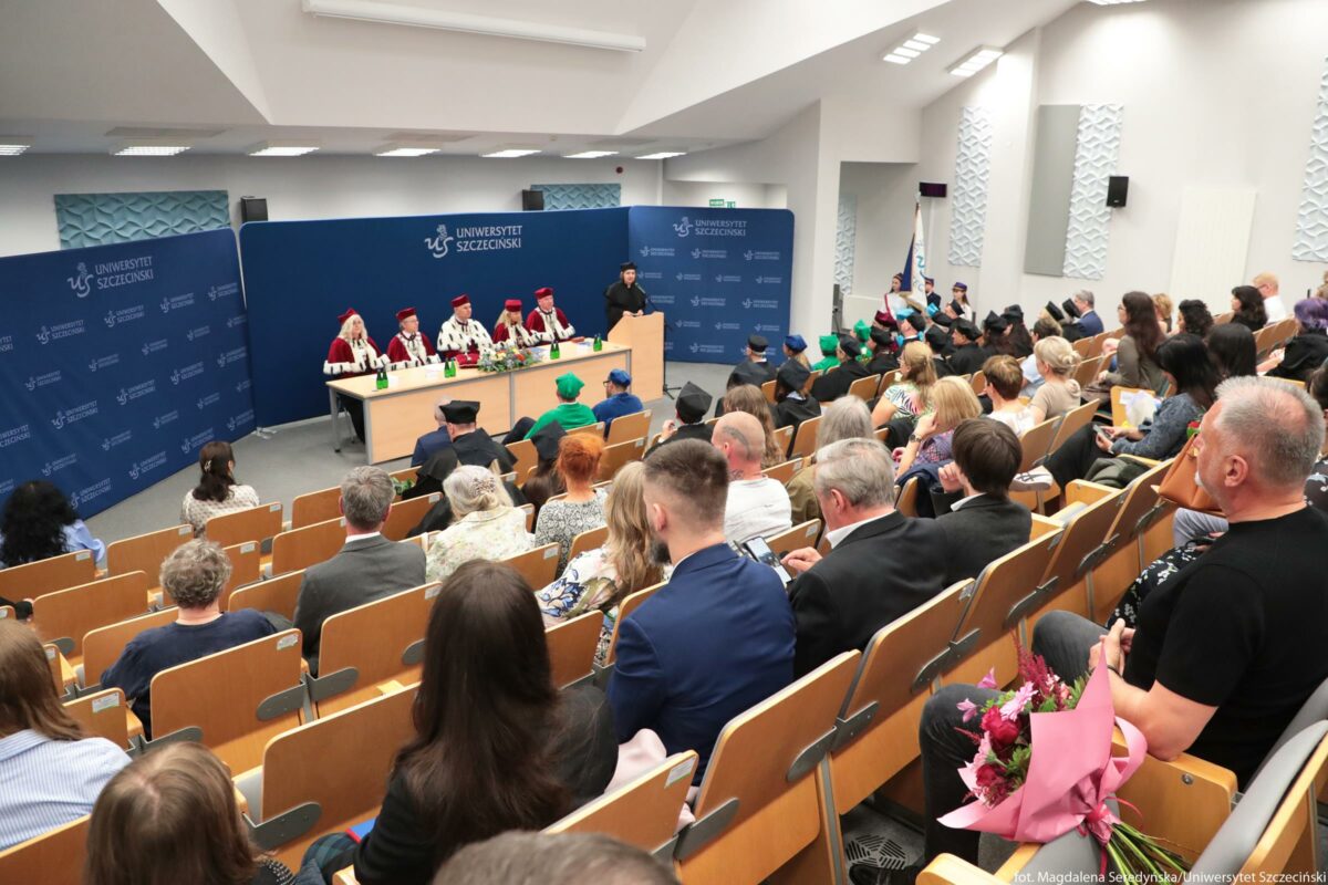 Uroczysta akademicka sala wykładowa z widownią i stołem prezydialnym na podwyższeniu z przodu. Na podium stoi kilka osób w tradycyjnych czerwono‑białych togach i biretach, prawdopodobnie władze uczelni, za długim stołem przykrytym jasnym blatem. Na stole stoją mikrofony, butelki z zielonymi etykietami oraz niewielkie dekoracje z kwiatów. Tło za stołem stanowi duża niebieska ścianka z powtarzającym się białym napisem „UNIWERSYTET SZCZECIŃSKI” i logo uczelni. Po prawej stronie przy mównicy przemawia osoba w ciemnym stroju. Przed podium widać rzędy drewnianych ławek z oparciami, wszystkie skierowane w stronę stołu i mównicy; miejsca są niemal całkowicie zajęte. Na widowni siedzą osoby w eleganckich ubraniach, część z nich ma na głowach birety w różnych kolorach, między innymi zielonym i niebieskim. W dolnym prawym rogu kadru jedna osoba trzyma bukiet kwiatów owinięty w różowy papier. Oświetlenie jest jasne, pochodzi z prostokątnych lamp sufitowych; ściany są jasne, a sala ma nowoczesny, uporządkowany wygląd.