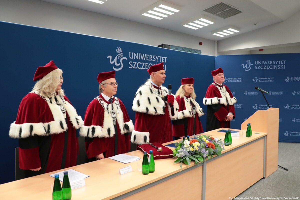 Pięć osób w uroczystych, czerwonych togach z białymi, futrzanymi obszyciami stoi za długim stołem. Wszystkie mają na głowach czerwone birety i srebrne łańcuchy z insygniami, co sugeruje skład władz uczelni lub senatu akademickiego. Osoba stojąca pośrodku trzyma mikrofon przy ustach, jakby przemawiała. Na stole leżą dokumenty, kilka zielonych szklanych butelek z wodą oraz bukiet kwiatów w odcieniach żółtym, białym, fioletowym i pomarańczowym. Na granatowej ścianie za nimi znajduje się biały napis „UNIWERSYTET SZCZECIŃSKI” oraz logo uczelni powtórzone w kilku miejscach. Po prawej stronie widoczny jest mównica z ustawionym mikrofonem. Scena odbywa się w jasnym, nowoczesnym pomieszczeniu z oświetleniem sufitowym.