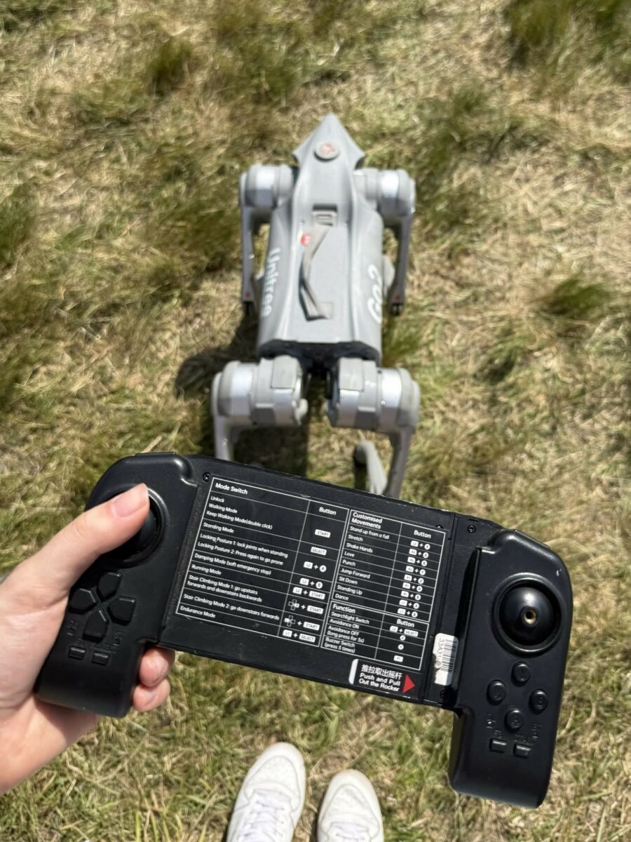 Czteronożny robot na trawie, widziany z góry. Robot jest szary, ma cztery grube nogi z przegubami i smukły korpus w kształcie wydłużonego sześciokąta. Na bokach korpusu widoczne są białe napisy „Unitree” i „GO2”, na górze jest prostokątny panel oraz okrągły element w przedniej części. Na pierwszym planie znajduje się czarny kontroler trzymany w obu dłoniach. Kontroler jest szeroki, po lewej stronie ma krzyżak kierunkowy i kilka małych przycisków, po prawej zaś wystaje duży, kulisty drążek sterujący (joystick) i rząd przycisków poniżej. Na środku kontrolera przyklejona jest duża, prostokątna etykieta z tabelą opisującą funkcje przycisków i tryby pracy. Napisy na tej etykiecie są zbyt małe i nieczytelne na tym zdjęciu, więc nie da się ich dokładnie odczytać. Pod kontrolerem w dolnej części kadru widać czubki białych sportowych butów osoby sterującej, stojącej na suchej, żółtawozielonej trawie. Robot stoi kilka kroków przed tą osobą, również na trawiastym podłożu.