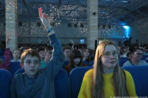Sala pełna siedzących ludzi, z chłopcem na pierwszym planie podnoszącym rękę. W pierwszym rzędzie widać chłopca o jasnej skórze w niebieskoszarej bluzie, z ręką uniesioną wysoko i dwoma wyprostowanymi palcami. Obok siedzi młoda kobieta o jasnej skórze i długich blond włosach, w okrągłych okularach i żółtym swetrze, patrząca przed siebie. W tle jest tłum publiczności na niebieskich fotelach oraz duża sala z kolumnami i punktowym oświetleniem tworzącym jasne plamki na suficie. W prawym dolnym rogu znajduje się mały podpis: „fot. Magdalena Seredyńska/Uniwersytet Szczeciński”.