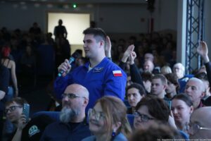 Mężczyzna stojący w auli wśród siedzącej publiczności, trzymający mikrofon i mówiący. Ma krótkie ciemne włosy i jasną skórę, ubrany jest w niebieski kombinezon z naszywkami; na prawym ramieniu widoczna jest flaga Polski, a na piersi emblemat z białym symbolem przypominającym ptaka. Wokół siedzi dużo osób; kilka ma podniesione ręce, część patrzy w stronę mówcy. Na dole po prawej jest podpis zdjęcia: „fot. Magdalena Seredyńska/Uniwersytet Szczeciński”.