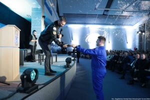 Aula z mężczyzną na podwyższeniu, który pochyla się i podaje coś mężczyźnie stojącemu niżej przy krawędzi sceny. Mężczyzna na scenie trzyma w drugiej ręce mikrofon i ma na sobie ciemną, błyszczącą kurtkę z naszywkami oraz ciemne spodnie; ma krótkie włosy i jasną skórę. Mężczyzna stojący przy scenie ma jasną skórę, krótkie ciemne włosy i jest ubrany w niebieski kombinezon; wyciąga obie ręce, żeby odebrać przekazywany przedmiot. W tle, po lewej stronie, stoi kobieta z mikrofonem, w okularach, w ciemnym stroju z jasną kamizelką. Po prawej stronie widać liczną publiczność siedzącą w rzędach. Na scenie stoją reflektory i sprzęt oświetleniowy; na ścianie za sceną jest jasna projekcja z abstrakcyjnym, białym wzorem. W prawym dolnym rogu jest podpis: „fot. Magdalena Seredyńska/Uniwersytet Szczeciński”.