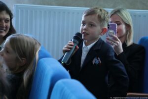 Chłopiec w szkolnym stroju trzymający mikrofon w sali z niebieskimi fotelami. Ma jasną skórę, krótkie jasnobrązowe włosy i białą koszulę pod ciemną marynarką; na marynarce jest naszywka z literami „SP” i małymi flagami. Siedzi wśród innych osób; za nim blond kobieta o jasnej skórze nagrywa telefonem, a po lewej widać uśmiechniętą kobietę. W tle jest biały grzejnik. W prawym dolnym rogu jest podpis: „fot. Magdalena Seredyńska/Uniwersytet Szczeciński”.