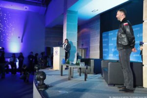 Scena w sali z niebiesko‑fioletowym oświetleniem. Po prawej stronie stoi dorosły mężczyzna o jasnej skórze, z krótkimi ciemnymi włosami i krótkim zarostem, ubrany w czarną skórzaną kurtkę z naszywką polskiej flagi na rękawie, ciemne spodnie i czarne buty; jest ustawiony bokiem i zwrócony w stronę mównicy. Na środku, przy mównicy, stoi dorosła kobieta o jasnej skórze i ciemnych włosach do ramion, w ciemnym stroju; mówi do mikrofonu. Za nią widać niebieskie tło z powtarzającym się napisem „UNIWERSYTET SZCZECIŃSKI” i logo. Na scenie stoją czarne kanapy oraz stolik z kilkoma zielonymi szklanymi butelkami i szklankami. Po lewej stronie, przy krawędzi sceny, znajduje się kilka osób i sprzęt fotograficzny/filmowy; na podłodze są też reflektory sceniczne. W prawym dolnym rogu widoczny jest podpis: „fot. Magdalena Seredyńska/Uniwersytet Szczeciński”.