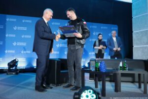 Dwóch mężczyzn na scenie podaje sobie ręce, a jeden z nich trzyma dużą, przezroczystą plakietę lub dyplom w ramie. Mężczyzna po lewej jest starszy, w granatowym garniturze, białej koszuli i krawacie. Mężczyzna po prawej jest młodszy, z krótkimi ciemnymi włosami i zarostem, ubrany w czarną skórzaną kurtkę z małą naszywką w barwach Polski na rękawie oraz w szare jeansy i ciemne buty. W tle znajduje się niebieska ścianka z powtarzającym się napisem „UNIWERSYTET SZCZECIŃSKI” i logo uczelni. Po prawej stronie stoją jeszcze dwie osoby: kobieta w okularach trzyma teczkę oraz mężczyzna w okularach w niebieskiej marynarce. Na scenie stoją ciemne fotele i niski stolik. Na stoliku są butelki (zielone i przezroczyste), szklanki oraz niebieski, dekoracyjny obiekt przypominający rzeźbę. Na dole po prawej widać podpis: „fot. Magdalena Seredyńska/Uniwersytet Szczeciński”.