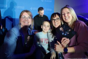 Grupa ludzi pozujących do zdjęcia na wydarzeniu z tłem w kolorze niebieskim z logo i napisem „UNIWERSYTET SZCZECIŃSKI”. Na pierwszym planie są trzy dorosłe kobiety i chłopiec, wszyscy uśmiechnięci i zwróceni w stronę aparatu. Kobiety mają jasną skórę; jedna po lewej ma duże, okrągłe okulary i jasnobrązowe włosy, środkowa ma ciemne włosy z grzywką i czarne okulary, a prawa ma długie blond włosy. Chłopiec na środku ma krótkie ciemne włosy i jasną skórę; ma na sobie szarą bluzę z napisem „NEW YORK”. Dwie kobiety trzymają zdjęcia przedstawiające mężczyznę w ciemnym stroju (wygląda jak kombinezon), a jedna z nich trzyma też przezroczystą butelkę. W tle, za nimi, stoi mężczyzna w czarnej bluzie, patrzący w dół, jakby podpisywał lub przygotowywał coś na stole. W prawym dolnym rogu widoczny jest podpis: „fot. Magdalena Seredyńska/Uniwersytet Szczeciński”.