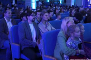 Publiczność siedząca w rzędach w auli o niebieskim oświetleniu. W pierwszym planie widać kilka osób siedzących obok siebie na niebieskich, tapicerowanych fotelach z drewnianymi podłokietnikami; większość patrzy w tym samym kierunku, jak na scenę poza kadrem. Najbliżej po prawej siedzi dorosła kobieta w okularach, a przed nią dwoje dzieci; jedno dziecko ma jasne włosy i bluzę we wzory. W środkowej części pierwszego planu siedzi mężczyzna z ciemnymi włosami, zarostem i okularami oraz kobieta z jasnymi włosami związanymi z tyłu, oboje wyglądają na skupionych. W tle widoczne są kolejne rzędy ludzi, w tym młodzież i dorośli. Na dole po prawej jest mały napis: „fot. Magdalena Seredyńska/Uniwersytet Szczeciński”.