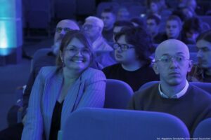 Grupa osób siedzących na widowni w sali, oświetlona niebieskim światłem. Na pierwszym planie po lewej stronie znajduje się uśmiechnięta kobieta w okularach, z krótkimi włosami, w jasnej marynarce w prążki. Po prawej stronie na pierwszym planie siedzi łysy mężczyzna w okularach, z poważnym wyrazem twarzy, ubrany w sweter i koszulę. Między nimi, nieco z tyłu, siedzi młoda osoba w okularach z ciemnymi włosami, patrząca w bok. W tle widać więcej osób na fotelach, część twarzy jest rozmyta. W prawym dolnym rogu jest drobny napis: „fot. Magdalena Seredyńska/Uniwersytet Szczeciński”.