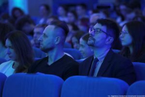 Grupa osób siedzących na widowni w auli, oświetlona niebieskim światłem. W pierwszym planie widać rzędy niebieskich, tapicerowanych foteli oraz kilka osób siedzących obok siebie. Najbardziej wyraźni są dwaj mężczyźni: jeden ma krótko ogolone boki głowy i dłuższe włosy na górze, drugi ma krótkie ciemne włosy, okulary, zarost i jest ubrany w marynarkę z koszulą. Po lewej stronie kadru siedzi kobieta z długimi włosami, patrząca w dół. W tle widać wielu innych uczestników, rozmytych, również siedzących i skierowanych w tę samą stronę. W prawym dolnym rogu jest mały podpis: „fot. Magdalena Seredyńska/Uniwersytet Szczeciński”.