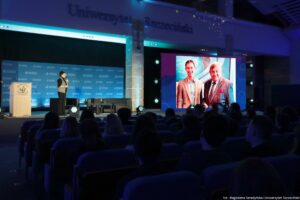 Aula podczas wydarzenia na Uniwersytecie Szczecińskim, z oświetloną sceną i publicznością na pierwszym planie. Na ścianie u góry widoczny jest napis „Uniwersytet Szczeciński”. Po lewej stronie stoi mównica z logo „US” i napisem „Uniwersytet Szczeciński”, a obok na scenie znajduje się osoba prowadząca w ciemnym stroju z jasną kamizelką. Tło sceny ma niebieską ściankę z powtarzającymi się logotypami uczelni. Po prawej stronie sceny widać duży ekran, na którym wyświetlone jest zdjęcie dwóch mężczyzn w marynarkach: młodszego po lewej i starszego po prawej. W dolnej części zdjęcia widać siedzącą publiczność w rzędach foteli. W prawym dolnym rogu jest podpis: „fot. Magdalena Seredyńska/Uniwersytet Szczeciński”.