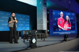 Scena w auli na uczelni z kobietą przemawiającą do mikrofonu oraz dużym ekranem po prawej stronie. Po lewej stronie stoi kobieta w średnim wieku o jasnej skórze, w okularach, z ciemnymi włosami związanymi z tyłu. Ma na sobie czarną bluzkę i spodnie oraz szarą kamizelkę. Trzyma mikrofon przy ustach i stoi obok czarnej kanapy. Na środku sceny stoją dwa niskie, czarne stoliki z kilkoma szklankami i zielonymi butelkami (prawdopodobnie woda). Z przodu na podłodze widać reflektory sceniczne. Tło stanowi granatowa ścianka z powtarzającym się napisem „UNIWERSYTET SZCZECIŃSKI” i logo „US”. Po prawej stronie jest duży ekran pokazujący zdjęcie dwóch osób w czerwonych koszulkach polo z naszywkami. Osoba po lewej ma krótkie siwe włosy i okulary, osoba po prawej ma długie ciemne kręcone włosy; obie mają jasną skórę. Trzymają czerwone pudełko. Na dole ekranu jest tekst: „Astrobiolodzy, astrofizycy prof. dr hab. Ewa Szuszkiewicz oraz dr hab. Franco Ferrari prof. US z Instytutu Fizyki US ich ‚kosmiczne’ odkrycie.” W prawym dolnym rogu zdjęcia jest podpis: „fot. Magdalena Seredyńska/Uniwersytet Szczeciński”.