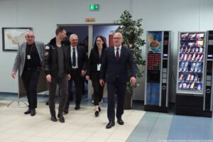 Grupa pięciu dorosłych osób idących korytarzem wewnątrz budynku. Z przodu po prawej idzie mężczyzna w granatowym garniturze, białej koszuli i ciemnym krawacie, w okularach. Po lewej idzie mężczyzna w szarej marynarce i czarnym ubraniu, z identyfikatorem na szyi. Obok niego idzie mężczyzna w ciemnej kurtce z naszywkami na rękawie. W tle w drzwiach widać kolejnego mężczyznę w garniturze oraz kobietę z długimi włosami, w ciemnym stroju i z identyfikatorem. Nad drzwiami jest zielony znak wyjścia ewakuacyjnego, a nad futryną tabliczka „110–117”. Po prawej stronie stoją dwa automaty: jeden z napojami kawowymi (grafika filiżanki) i drugi z przekąskami i napojami. Obok automatów stoi wysoka roślina w donicy. Na dole po prawej widoczny jest podpis: „fot. Magdalena Seredyńska / Uniwersytet Szczeciński”.