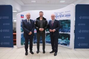 Trzech mężczyzn stojących w pomieszczeniu przed ścianką promocyjną. Mężczyzna po lewej jest starszy, ma krótkie siwe włosy, ciemny garnitur z białą koszulą i ciemnym krawatem. Mężczyzna w środku jest młodszy, ma krótkie ciemne włosy i krótki zarost, czarną skórzaną kurtkę na ciemnym ubraniu oraz ciemne spodnie. Mężczyzna po prawej jest starszy, łysy, ma okulary, ciemny garnitur z białą koszulą i bordowym krawatem. W tle są dwie granatowe ścianki z powtarzającym się napisem „UNIWERSYTET SZCZECIŃSKI” i logo. Na centralnej białej ściance widoczny jest napis „Odkryj na dobre _ Szczecin” oraz duży napis „Szczecin 2050” z mniejszym angielskim tekstem „floating garden project”. Jest też małe logo „Szczecin” oraz grafiki miasta w odcieniach niebieskiego. Podłoga jest z jasnych płytek, sufit z panelami i lampami biurowymi. W prawym dolnym rogu widoczny podpis: „fot. Magdalena Seredyńska/Uniwersytet Szczeciński”.