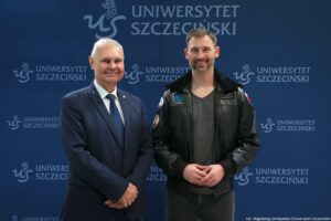 Dwóch mężczyzn stojących obok siebie na tle granatowej ścianki z logo i napisem „UNIWERSYTET SZCZECIŃSKI”. W tle powtarza się tekst „UNIWERSYTET SZCZECIŃSKI” oraz znak „US”. Po lewej stoi starszy mężczyzna o jasnej skórze, krótkich siwych włosach, w ciemnym garniturze z białą koszulą i ciemnym krawatem; ma złączone dłonie z przodu i uśmiecha się. Po prawej stoi młodszy mężczyzna o jasnej skórze, krótkich brązowych włosach i krótkim zaroście; ma na sobie czarną skórzaną kurtkę z naszywkami i szary T-shirt, dłonie trzyma z przodu i uśmiecha się. Na dole po prawej jest drobny podpis: „fot. Magdalena Seredyńska/Uniwersytet Szczeciński”.