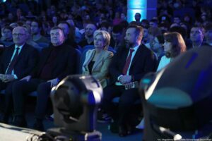 Rząd osób siedzących w pierwszym rzędzie w auli, oświetlonych niebieskim światłem. Widać pięć osób na pierwszym planie: trzech mężczyzn w garniturach (jeden łysy w okularach, jeden w czarnym golfie, jeden z brodą i czerwonym krawatem) oraz dwie kobiety (jedna z krótkimi blond włosami w jasnej marynarce, druga z brązowymi włosami w jasnej bluzce). Wszyscy siedzą na niebieskich fotelach i mają neutralne, skupione wyrazy twarzy, skierowane w stronę sceny poza kadrem. W tle znajduje się duża publiczność na widowni, również w niebieskawym oświetleniu. Na dole kadru są rozmyte elementy sprzętu scenicznego lub oświetleniowego. W prawym dolnym rogu widoczny jest napis: „fot. Magdalena Seredyńska/Uniwersytet Szczeciński”.