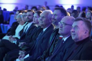 Rząd siedzących osób na widowni podczas wydarzenia w sali, oświetlonych niebiesko-fioletowym światłem. W pierwszym planie widać kilku mężczyzn w garniturach i jednego mężczyznę w ciemnej kurtce z małą przypinką w barwach biało-czerwonych; większość ma jasną skórę. Po lewej stronie rzędu siedzi starsza kobieta z krótkimi jasnymi włosami i okularami, ubrana na jasno. W tle znajduje się większa publiczność, rozmyta. W prawym dolnym rogu widoczny jest napis: „fot. Magdalena Seredyńska/Uniwersytet Szczeciński”.
