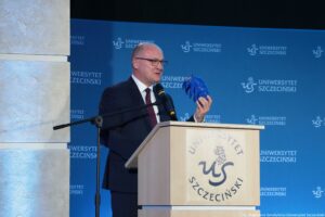 Mężczyzna w garniturze stojący przy mównicy z logo „Uniwersytet Szczeciński”. Ma jasną skórę, jest łysy i nosi okulary. Przemawia do mikrofonu i trzyma w dłoni niebieski przedmiot przypominający model dłoni lub rękawicę. W tle znajduje się niebieska ścianka z powtarzającym się napisem „UNIWERSYTET SZCZECIŃSKI” i logo „US”. Na dole po prawej widoczny jest mały podpis: „fot. Magdalena Seredyńska/Uniwersytet Szczeciński”.