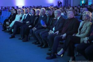 Rząd siedzących osób na widowni w auli, oświetlonych niebieskim światłem. W pierwszym rzędzie siedzą kobiety i mężczyźni w eleganckich strojach: garniturach, marynarkach i ciemnych sukienkach; po prawej stronie widoczna kobieta w jasnej marynarce. W środkowej części pierwszego rzędu mężczyzna trzyma w dłoniach niebieski, żebrowany przedmiot. Za nimi widać wiele kolejnych osób siedzących na niebieskich fotelach. W prawym dolnym rogu jest podpis: „fot. Magdalena Seredyńska/Uniwersytet Szczeciński”.