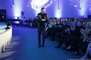 Mężczyzna idący przejściem obok sceny w auli, w czarnej kurtce i ciemnych spodniach, trzymający w dłoni niebieski przedmiot. Ma jasną skórę i krótko przycięte włosy oraz zarost. Po prawej stronie siedzi liczna publiczność na fotelach; wiele osób klaszcze. Oświetlenie jest niebiesko-fioletowe, a na ścianie w tle widać jasną projekcję przypominającą galaktykę lub spiralę. Po lewej stronie znajduje się fragment sceny z reflektorami i stojakiem/mikrofonem. W prawym dolnym rogu widoczny jest mały podpis: „fot. Magdalena Seredyńska/Uniwersytet Szczeciński”.