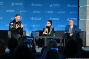 Panel dyskusyjny na scenie z trzema osobami siedzącymi na czarnych fotelach, na tle niebieskiej ścianki z powtarzającym się napisem „UNIWERSYTET SZCZECIŃSKI” i logo uczelni. Po lewej siedzi mężczyzna o jasnej skórze, z krótkimi ciemnymi włosami i zarostem, w czarnej skórzanej kurtce z naszywkami; trzyma mikrofon i gestykuluje podczas mówienia. Pośrodku siedzi kobieta o jasnej skórze, w okularach, z ciemnymi włosami upiętymi w kok; trzyma mikrofon i ma na kolanach notatki. Po prawej siedzi mężczyzna o jasnej skórze w okularach, w niebieskiej marynarce i szarej kamizelce; trzyma notatnik lub teczkę i patrzy w stronę rozmówców. Przed nimi stoją niskie czarne stoliki z kilkoma zielonymi butelkami, szklankami oraz niebieskim dekoracyjnym obiektem. Na dole kadru widać publiczność w półcieniu; ktoś unosi telefon, nagrywając lub robiąc zdjęcie. W prawym dolnym rogu jest podpis: „fot. Magdalena Seredyńska/Uniwersytet Szczeciński”.