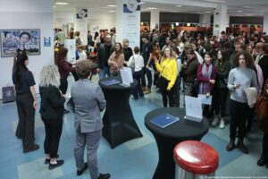 Zatłoczone wnętrze budynku z dużą grupą ludzi stojących w kolejce lub tłumie. Na pierwszym planie stoją trzy wysokie, okrągłe stoliki koktajlowe z czarnymi pokrowcami; na dwóch z nich leżą niebieskie teczki lub foldery, a na jednym stoi przezroczysta podstawka na kartkę. Z prawej na dole widać fragment okrągłego, czerwonego siedziska. W tle jest bardzo dużo osób (głównie młodych dorosłych) w kurtkach i swetrach; część trzyma kartki lub dokumenty. Pośrodku, bliżej prawej strony, wyróżnia się osoba w jaskrawożółtym swetrze. W głębi widać białe filary i podwieszone banery z okrągłym logo „US” oraz napisem „Uniwersytet Szczeciński”. Na ścianie po lewej jest obraz/plakat z twarzą oraz kilka małych tabliczek informacyjnych. Na dole po prawej jest podpis: „fot. Magdalena Seredyńska/Uniwersytet Szczeciński”.