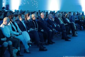 Aula z dużą publicznością siedzącą w rzędach, oświetlona niebieskim światłem. Na pierwszym planie widać rząd kilkunastu osób siedzących na krzesłach, zwróconych w lewą stronę, jak podczas wykładu lub uroczystości. Większość osób ma jasną karnację. Ubrania są w większości formalne: garnitury, marynarki, eleganckie sukienki i swetry; kilka osób ma okulary. Po prawej stronie widać kolejne rzędy siedzących ludzi, a z tyłu sali tłum w półmroku. Na ścianie w tle są jasne plamy światła i drobne refleksy. W prawym dolnym rogu jest podpis: „fot. Magdalena Seredyńska/Uniwersytet Szczeciński”.