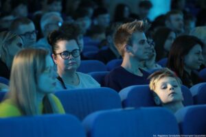 Aula z publicznością siedzącą w niebieskich, tapicerowanych fotelach. W kadrze dominuje kilka osób na pierwszych planach: kobieta w średnim wieku o jasnej skórze, w czarnych okularach, z ciemnymi włosami upiętymi w kok, w jasnoszarej bluzce; obok niej młody mężczyzna o jasnej skórze, z krótkimi jasnobrązowymi włosami, w ciemnej koszulce, zwrócony profilem. Na dole po prawej widoczne jest dziecko o jasnej skórze i jasnych włosach, odchylone do tyłu na fotelu. Po lewej na pierwszym planie znajduje się rozmyta postać kobiety z długimi blond włosami. Tło wypełnia większa grupa osób, częściowo nieostra, oświetlona chłodnym, niebieskawym światłem. W prawym dolnym rogu widoczny jest mały podpis: „fot. Magdalena Seredyńska/Uniwersytet Szczeciński”.