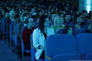 Aula wypełniona publicznością siedzącą w rzędach niebieskich foteli. W kadrze dominują osoby siedzące po lewej stronie, głównie dorośli i kilka dzieci, wszyscy zwróceni w tym samym kierunku, jak podczas wykładu lub wydarzenia. Oświetlenie ma chłodny, niebieskawy odcień. Na pierwszym planie siedzi kobieta z długimi ciemnymi włosami w białej marynarce, obok niej inne osoby w okularach; w tle widać gęsty tłum na kolejnych rzędach. W prawym dolnym rogu jest podpis: „fot. Magdalena Seredyńska/Uniwersytet Szczeciński”.