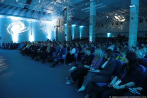 Aula wypełniona publicznością siedzącą w rzędach niebieskich foteli. Oświetlenie jest chłodne, niebieskie, a na ścianach i suficie widać liczne świetlne punkty przypominające projekcję gwiazd lub refleksy. Po lewej stronie na ścianie wyświetlony jest jasny, spiralny symbol przypominający galaktykę. W pierwszych rzędach siedzą głównie dorośli w formalnych strojach (garnitury, marynarki), a dalej widoczny jest tłum osób w mniej formalnym ubiorze. W tle stoją wysokie filary i elementy oświetlenia scenicznego. W prawym dolnym rogu widoczny jest napis: „fot. Magdalena Seredyńska/Uniwersytet Szczeciński”.