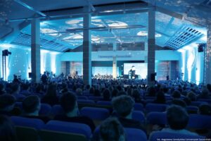 Aula wypełniona publicznością, oświetlona na niebiesko, z widokiem na scenę na końcu sali. Na suficie i ścianach widać projekcje świetlne przypominające gwiazdy i planety. Z przodu znajduje się scena z panelem dyskusyjnym i dużym ekranem, na którym wyświetlany jest mężczyzna mówiący do mikrofonu. Nad sceną widoczny napis „Uniwersytet Szczeciński”. W sali stoją wysokie filary, a po bokach widać sprzęt nagraniowy i kamery. W prawym dolnym rogu jest podpis: „fot. Magdalena Seredyńska/Uniwersytet Szczeciński”.