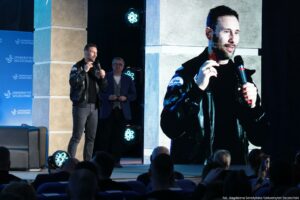 Scena konferencyjna z mężczyzną przemawiającym do publiczności i dużym ekranem z jego zbliżeniem. Mężczyzna ma krótkie ciemne włosy i krótki zarost, trzyma mikrofon w prawej dłoni i gestykuluje drugą ręką. Jest ubrany w czarną skórzaną kurtkę i ciemną koszulkę. Na rękawie kurtki widać naszywkę z napisem „Ignis”. Po lewej stronie jest niebieskie tło z powtarzającym się białym napisem „UNIWERSYTET SZCZECIŃSKI” i logo uczelni, a obok stoi szary fotel. Za mówcą stoi drugi mężczyzna w okularach, w granatowej marynarce, obserwujący wystąpienie. Na pierwszym planie widać głowy osób z publiczności. W prawym dolnym rogu jest podpis: „fot. Magdalena Seredyńska/Uniwersytet Szczeciński”.