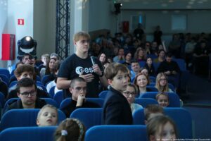 Aula wypełniona publicznością siedzącą w niebieskich fotelach. Na środku kadru stoi młody mężczyzna o jasnych włosach i jasnej skórze, w okularach, w czarnym t-shircie z białym napisem, trzyma mikrofon i wygląda, jakby zadawał pytanie lub zabierał głos. Na pierwszym planie widać chłopca o jasnej skórze w ciemnej marynarce, odwróconego bokiem w stronę aparatu. W tle siedzą dorośli i dzieci, część osób patrzy w stronę mówiącego. Po lewej stronie widać elementy wyposażenia sali: czarną lampę sceniczną oraz czerwone skrzynki na ścianie. W prawym dolnym rogu jest podpis fotograficzny: „fot. Magdalena Seredyńska/Uniwersytet Szczeciński”.