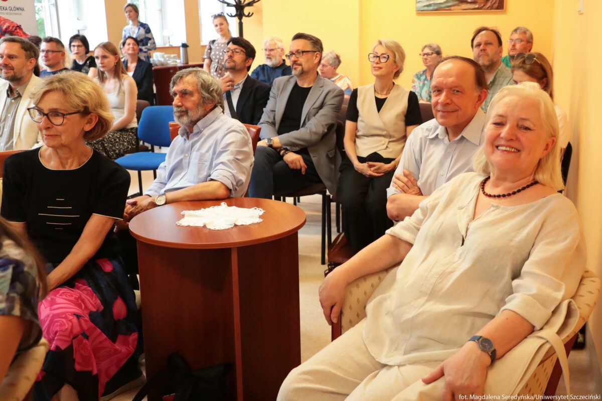 Grupa ludzi siedząca w niewielkiej, jasno oświetlonej sali. Na pierwszym planie, po prawej stronie, siedzi starsza kobieta o jasnych, siwych włosach do ramion, w jasnej, lnianej bluzce i spodniach, z naszyjnikiem z ciemnych koralików i zegarkiem na nadgarstku; uśmiecha się szeroko w kierunku aparatu. Obok niej, nieco z tyłu, siedzi mężczyzna w jasnoniebieskiej koszuli z podwiniętymi rękawami, z rękami skrzyżowanymi na piersi i lekkim uśmiechem. Po lewej stronie, na przodzie, znajduje się kobieta o krótkich, blond włosach, w czarnej bluzce i długiej spódnicy w duże różowo-fioletowe kwiaty; patrzy w stronę przodu sali. Przy niej stoi niski, okrągły stolik z ciemnego drewna, na którym leży biała szydełkowa serwetka. Za tym stolikiem siedzi starszy mężczyzna z siwymi, lekko potarganymi włosami i brodą, w jasnoniebieskiej koszuli z podwiniętymi rękawami, z rękami opartymi na kolanach. W kilku rzędach dalej widać kilkanaście osób – kobiety i mężczyzn w średnim i starszym wieku – w eleganckich, ale niezbyt formalnych ubraniach: marynarkach, koszulach, sukienkach i bluzkach. Twarze większości osób są pogodne, część się uśmiecha, część przygląda się czemuś poza kadrem, jakby słuchała wystąpienia. Krzesła są proste, konferencyjne, w różnych kolorach, głównie brązowych i niebieskich. Ściany sali są w ciepłym, żółtym kolorze, w tle wiszą obrazy i stoją stoliki z filiżankami, dzbankiem i innymi drobnymi przedmiotami, co sugeruje spotkanie towarzyskie lub kulturalne w przyjaznej atmosferze.