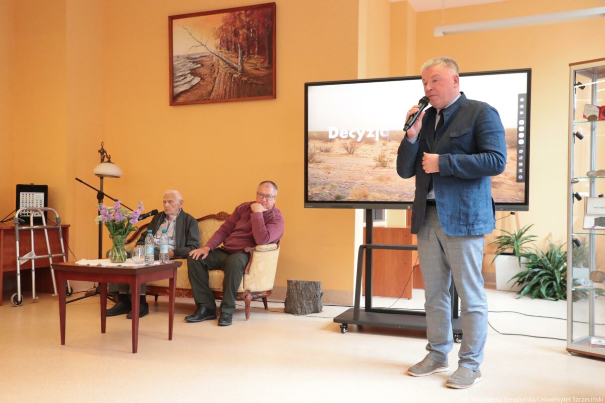 Spotkanie w jasnym pomieszczeniu z trzema mężczyznami i dużym ekranem. Po prawej stronie stoi mężczyzna w średnim wieku, o jasnych, krótko ostrzyżonych włosach. Ma na sobie niebieską marynarkę, jasnoszarą koszulę, krawat i szare spodnie w drobną kratkę oraz jasne buty. Trzyma w dłoni mikrofon i mówi, zwrócony lekko w stronę siedzących. Za nim stoi duży ekran na kółkach; na ekranie widoczny jest krajobraz pustynny, a na środku wyświetla się białe słowo „Decyzja”. Po lewej stronie zdjęcia siedzi dwóch starszych mężczyzn na kremowej, stylizowanej kanapie z drewnianymi bokami. Mężczyzna po lewej jest bardzo szczupły, siwowłosy, w ciemnych spodniach, jasnej koszuli i ciemnej marynarce; przed nim stoi mikrofon na statywie i kilka butelek wody na stole. Mężczyzna po prawej ma siwe włosy, jest ubrany w bordowy sweter i ciemne spodnie, siedzi wygodnie, podpierając policzek dłonią i patrzy w stronę mówiącego. Przed kanapą stoi mały drewniany stolik z przezroczystym wazonem z fioletowymi i jasnoróżowymi kwiatami oraz kilkoma plastikowymi butelkami wody i kubkami. Z lewej strony widać balkoniki rehabilitacyjne obok stolika pomocniczego, a nad kanapą wisi obraz przedstawiający jesienny las i wodę przy brzegu. Ściany są w ciepłym, jasnopomarańczowym kolorze. W głębi, po prawej, znajduje się przeszklona gablotka oraz kilka doniczkowych roślin. Jeśli chcesz, mogę opisać bardziej szczegółowo osoby, ich mimikę albo elementy wyposażenia sali.