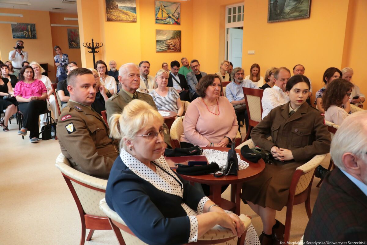 Sala z publicznością podczas spotkania lub uroczystości. Na pierwszym planie siedzi kilka osób przy małych, okrągłych stolikach z koronkowymi serwetkami i damskimi torebkami oraz okularami. Najbliżej znajduje się kobieta o jasnych blond włosach upiętych z tyłu, w granatowym żakiecie z białą, grochowaną klapą i mankietami; ma jasną skórę i okulary, patrzy w stronę prowadzącego wydarzenie. Po jej lewej stronie siedzi młody mężczyzna w mundurze wojskowym w kolorze oliwkowo‑brązowym, z naszywką „POLAND” na ramieniu i herbem w kształcie tarczy. Obok, nieco dalej, siedzi starszy mężczyzna o siwych włosach i jasnej skórze, również w eleganckim mundurze wojskowym, z wyprostowaną postawą. Z prawej strony kadru, przy tym samym stoliku, siedzi młoda kobieta o bardzo jasnej skórze, z ciemnymi, gładko zaczesanymi włosami związanymi z tyłu. Ma na sobie długi, brązowy mundur z dużymi kieszeniami i zapięciem pod szyję, trzyma dłonie splecione na kolanach. Za nimi, w kilku rzędach, siedzi większa grupa osób – kobiety i mężczyźni w różnym wieku, w ubraniach od eleganckich po bardziej codzienne, jak koszule, swetry czy bluzki. Wśród nich widać m.in. kobietę w różowej, falbaniastej bluzce, kilka osób w białych koszulach, mężczyzn w marynarkach oraz osoby z brodami i okularami. Wszyscy zwróceni są w jednym kierunku, wyglądają na skupionych i zasłuchanych. Ściany sali są w ciepłym, morelowo‑żółtym kolorze i wiszą na nich obrazy przedstawiające pejzaże i żaglówki. Na środku, przy słupie, stoi pusty stojak na płaszcze. W głębi, przy ścianie, jedna osoba fotografuje wydarzenie aparatem, a obok stoi kobieta w sukience patrząca na zgromadzonych. W tle widać też otwarte drzwi prowadzące do innego, jasnego pomieszczenia.