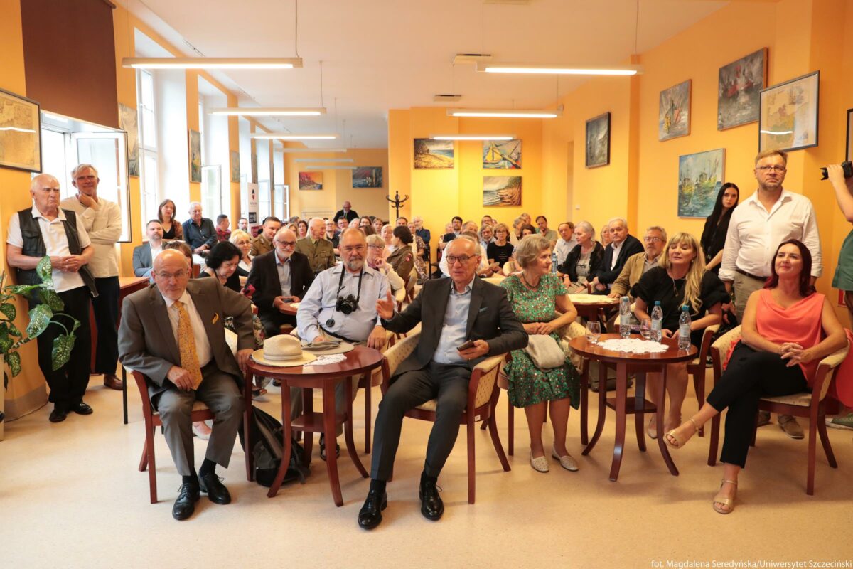Sala wypełniona ludźmi podczas spotkania lub uroczystości. Pomieszczenie ma żółtopomarańczowe ściany, jest długie, z rzędem dużych okien po lewej stronie, przez które wpada jasne dzienne światło. Na ścianach wiszą kolorowe obrazy, głównie pejzaże i sceny z żaglówkami. W całym kadrze siedzi i stoi kilkadziesiąt osób, większość w średnim i starszym wieku, wszystkie zwrócone twarzami w stronę aparatu, jakby pozowały do wspólnego zdjęcia lub słuchały kogoś znajdującego się tuż przed fotografującym. Uczestnicy są elegancko ubrani – mężczyźni głównie w garniturach lub koszulach, kobiety w sukienkach lub zestawach wizytowych. Widoczne są różne kolory włosów i odcienie skóry, ogólne wrażenie to zróżnicowana, ale dość formalna grupa. Na pierwszym planie stoją małe okrągłe stoliki z ciemnego drewna, otoczone krzesłami i fotelikami. Przy trzech najbliższych stolikach siedzi kilka osób wyróżniających się w kadrze: po lewej starszy mężczyzna w szarym garniturze z żółtym krawatem, obok niego inny starszy mężczyzna w jasnej koszuli z aparatem fotograficznym zawieszonym na szyi oraz mężczyzna w ciemnym garniturze z telefonem w dłoni, obok którego siedzi kobieta w zielonej sukience w kwiaty. Przy kolejnym stoliku po prawej siedzą dwie kobiety; na blacie stoją plastikowe butelki z wodą i kubki. W głębi widać gęsto ustawione krzesła zajęte przez publiczność; niektórzy stoją pod ścianami między oknami, inni opierają się o ścianę po prawej stronie. W tle na środku stoi wieszak na płaszcze. Oświetlenie zapewniają podłużne lampy sufitowe zawieszone w jednym rzędzie wzdłuż całej sali. Atmosfera na zdjęciu wydaje się spokojna i skupiona, jak podczas odczytu, spotkania autorskiego albo akademickiej uroczystości.
