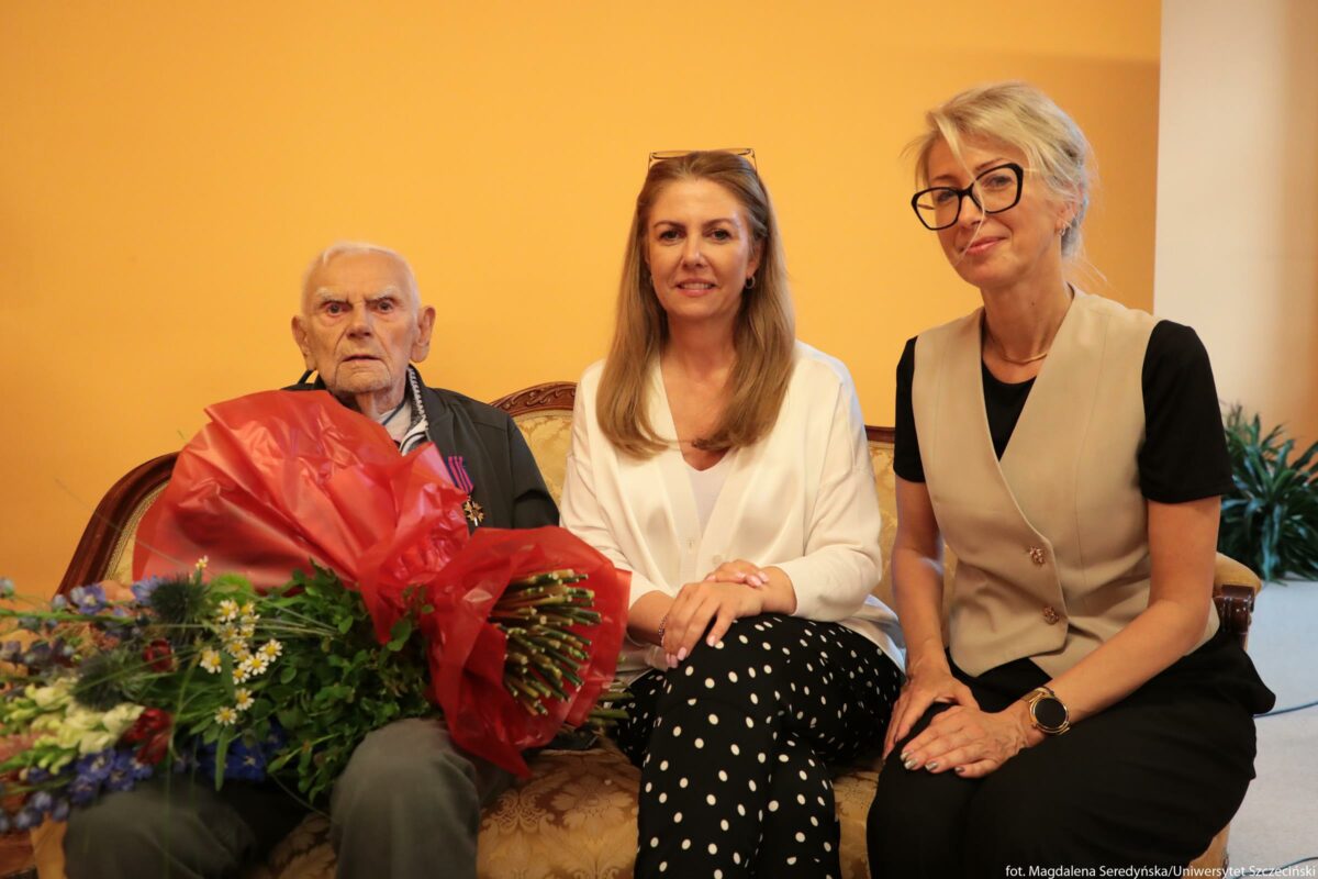 Trzy osoby siedzące obok siebie na kanapie na tle gładkiej, żółtopomarańczowej ściany. Po lewej stronie siedzi bardzo starszy mężczyzna o jasnej karnacji i krótkich, białych włosach. Ma poważny wyraz twarzy, lekko rozchylone usta i patrzy na wprost. Ubrany jest w ciemnozieloną kurtkę na zamek, pod którą widać biały kołnierzyk koszuli lub bluzy. Na piersi ma przypięte odznaczenia. Na kolanach trzyma bardzo duży bukiet kwiatów owinięty czerwoną, błyszczącą folią; z prawej strony wystaje wiele zielonych łodyg i liści, z lewej różnokolorowe kwiaty. W środku siedzi kobieta o jasnej karnacji i długich, prostych, blond włosach do ramion. Ubrana jest w białą, luźną bluzkę z dekoltem w serek i ciemne spodnie w białe grochy. Dłonie ma złożone na kolanach. Na głowie, nad czołem, ma założone okulary przeciwsłoneczne lub korekcyjne. Uśmiecha się lekko do obiektywu. Po prawej stronie siedzi druga kobieta o jasnej karnacji i jasnych, upiętych włosach z pasmem opadającym na czoło. Nosi duże, czarne okulary w grubych oprawkach, czarną koszulkę i beżową kamizelkę zapinaną na dwa guziki, a także czarne spodnie. Na lewym nadgarstku ma zegarek z dużą, okrągłą tarczą. Dłonie trzyma złożone na kolanach i uśmiecha się lekko. W tle, po prawej stronie, znajduje się niewielka roślina doniczkowa na podłodze. Całe ujęcie sprawia wrażenie uroczystego spotkania lub wizyty, prawdopodobnie z okazji uhonorowania starszego mężczyzny.