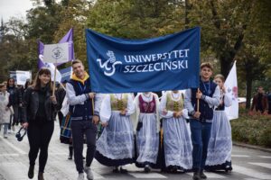 Uśmiechnięci dwaj studenci trzymają z dwóch stron granatowy transparent z białym napisem Uniwersytet Szczeciński. Po lewej studentka trzymająca mniejszy biały transparent z logiem US. Za nimi ubrane w stroje ludowe członkinie zespołu folkowego (twarze zakryte przez transparent). W tle inni uczestnicy i zielone drzewa.