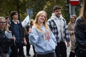 Na pierwszym planie studentka w błękitniej bluzie z postaciami z bajki o Kubusiu Puchatku trzyma małą białą flagę z granatowym logiem Uniwersytetu Szczecińskiego. Po lewej stronie (lekko w tle) druga studentka z taką samą flagą. W tle zielone drzewa i sygnalizacja świetlna oraz pozostali uczestnicy Spacerku na uniwerek.