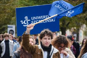 Student machający granatową flagą z białym napisem Uniwersytet Szczeciński. Za nim studentka trzymająca taki sam transparent. W tle młodzież – uczestnicy Spacerku na uniwerek.