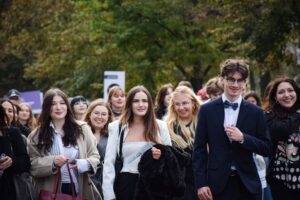 Grupa studentek – uczestniczek Spacerku na uniwerek. Po prawej student w garniturze, białej koszuli i muszce. W tle młodzież – pozostali uczestnicy wydarzenia.