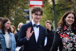 Na pierwszym planie machający student w garniturze, białej koszuli i muszce, obok (po prawej) studentka w bluzie z kolorowymi Atomówkami z bajki. W tle młodzież – uczestnicy Spacerku na uniwerek.