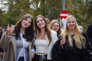 Na pierwszym planie cztery studentki – uczestniczki Spacerku na uniwerek. Pierwsza z prawej pokazuje znak Victorii, następna trzyma białą flagę z granatowym logiem Uniwersytetu Szczecińskiego. W tle zielone drzewa.