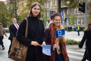 Na pierwszym planie dwie studentki trzymające granatowe flagi z logiem Spacerku na uniwerek. W tle po prawej kamienice, sygnalizacja świetlna i policjant kierujący ruchem.
